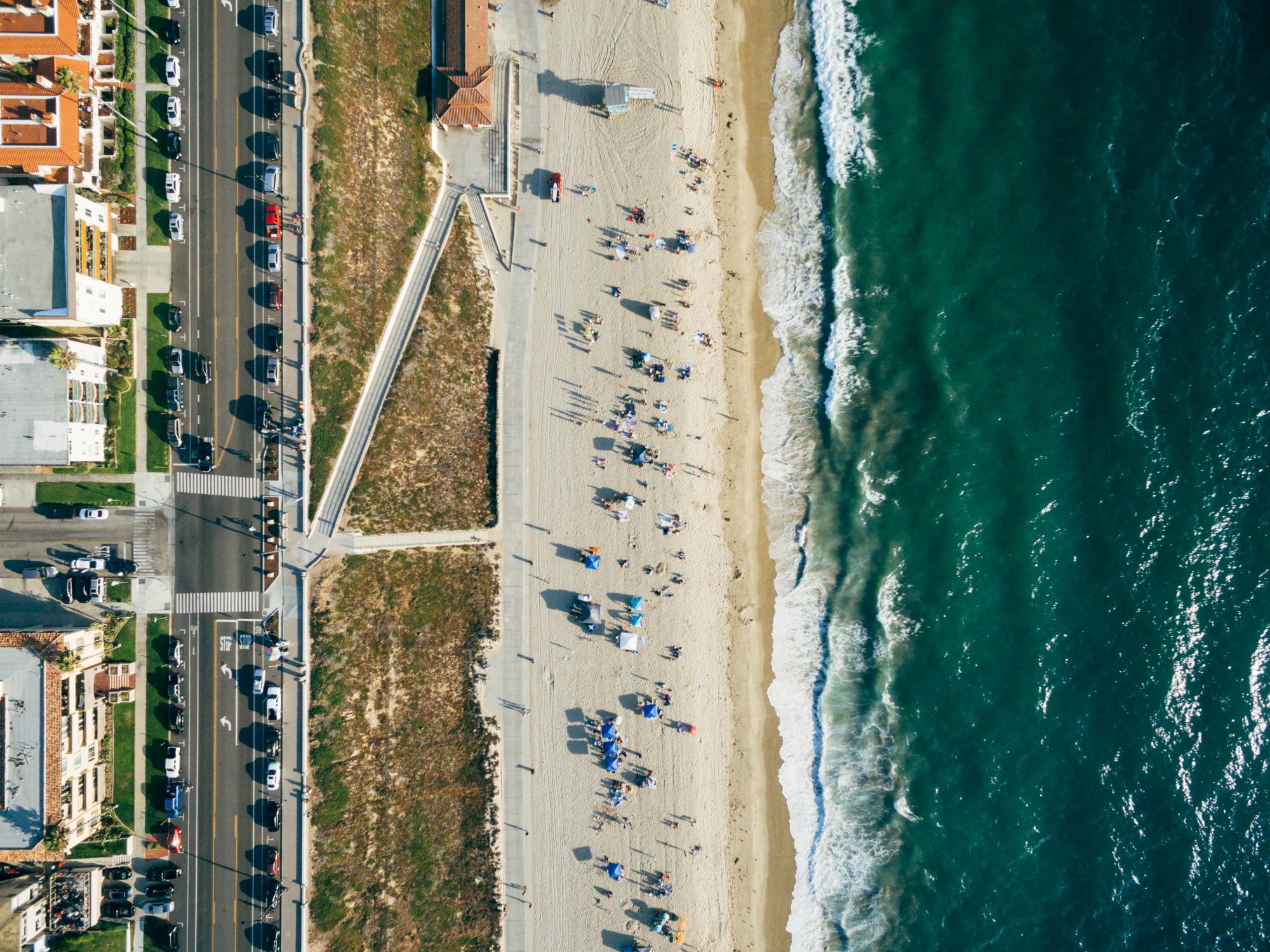 People sunbathing and strolling on a sandy beach beside turquoise waves, with a boardwalk and buildings lining the shore. ending the sentence.