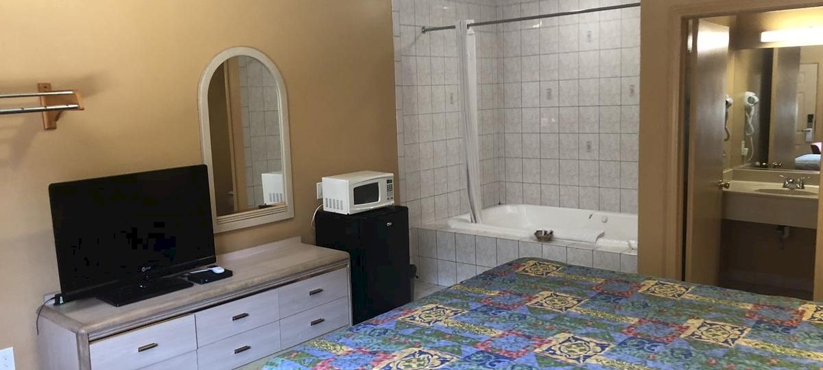 A cozy hotel room with a bed, dresser, TV, and a tiled bathroom area; quilted bedspread and a small safe beside a microwave.