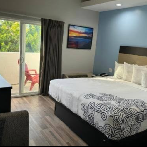 A hotel room with a large bed, blue accent wall, balcony door, and a balcony chair outside; desk and a framed landscape picture.