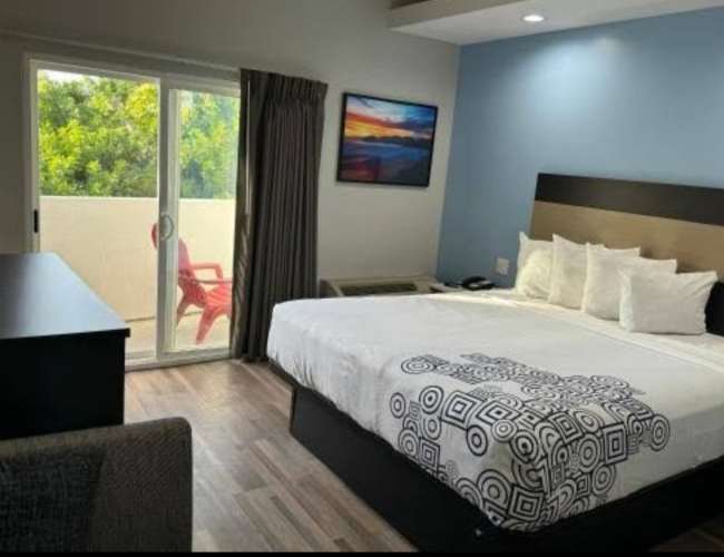 A hotel room with a large bed, blue accent wall, balcony door, and a balcony chair outside; desk and a framed landscape picture.
