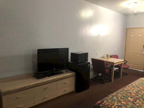 A simple hotel room setup with a TV on a low dresser, a mini-fridge and microwave, plus a small table and chairs near the wall, and a bed in the foreground.