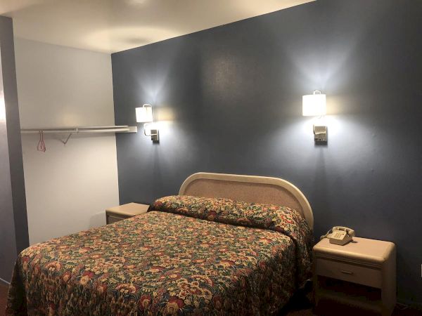 A hotel room with a double bed, dark blue accent wall, two wall sconces, a small nightstand with a phone, and an open closet area.