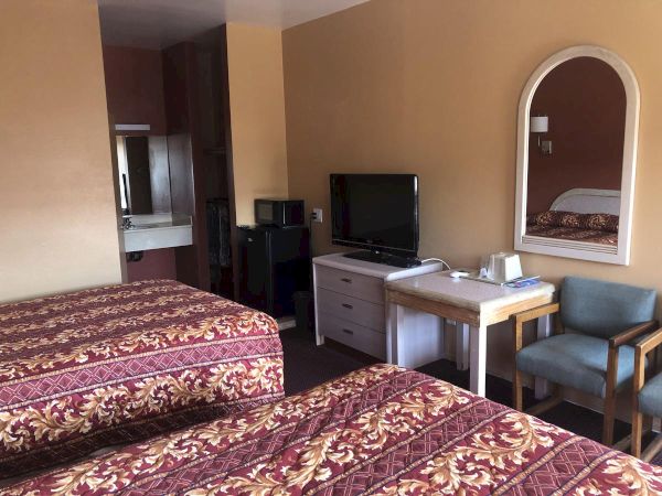 Two double beds with red-gold patterned bedspreads, a desk with a TV, small dresser, and a mirror on a beige wall in a hotel room.