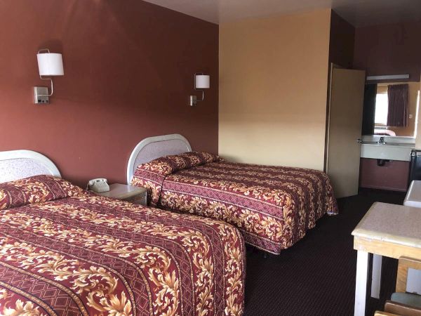Two double beds in a motel room with maroon and gold bedspreads, a small nightstand, wall lamps, and a kitchenette area in the back.