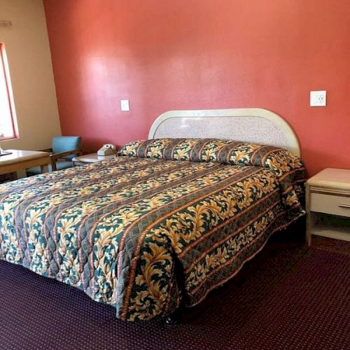 A hotel room with a large bed, red accent wall, patterned bedspread, two white nightstands, a small table and chair in the corner, and a carpeted floor.