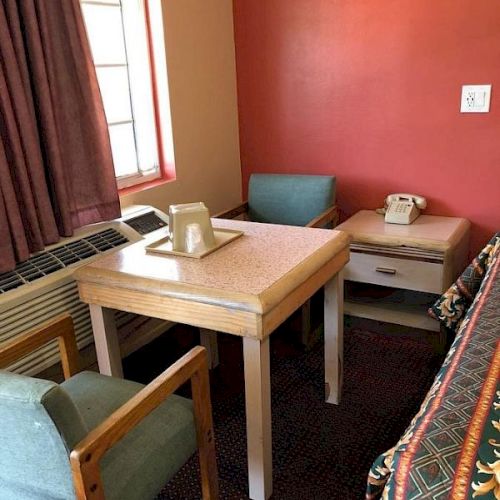 A small, cozy corner with a wooden table, two chairs, a sofa bed, a window with curtains, and a wall-mounted red accent wall with a telephone on a side table.