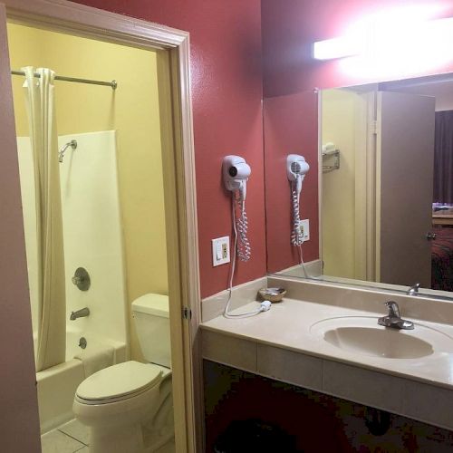 A hotel bathroom with a single sink, large mirror, wall-mounted hairdryer, and a separate tub/shower combo visible through the open door.