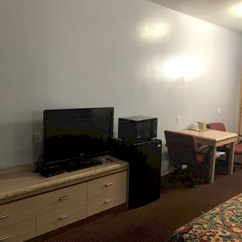 A basic hotel room or suite with a TV on a low cabinet, a small desk and chair, a printer or mini fridge, and a bed with a patterned bedspread.